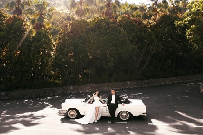 Couple sitting on a white classic car under palm trees