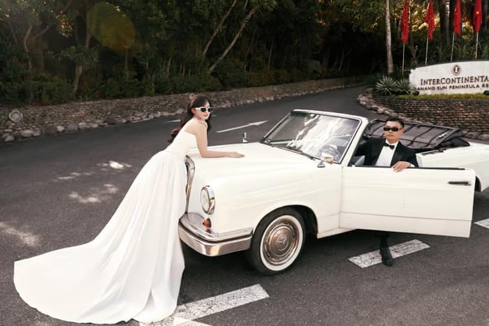 Bride leaning on a white classic car with groom seated inside